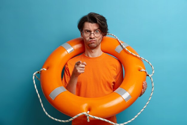 El rescatador hombre estricto disgustado grave le señala y advierte sobre el peligro en el agua, posa con un salvavidas, trabaja en una playa tropical, vestido con una camiseta naranja, listo para la salvación de la persona que se hunde