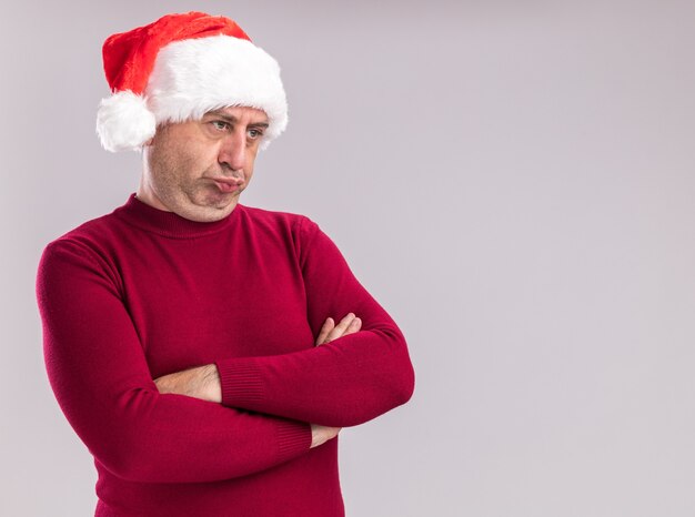 Resentido hombre de mediana edad vestido con gorro de Papá Noel de Navidad mirando a un lado con los brazos cruzados de pie sobre la pared blanca