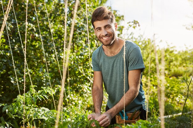 Respetuoso del medio ambiente. Concepto de estilo de vida saludable. Retrato al aire libre de joven agricultor caucásico barbudo atractivo sonriendo a puerta cerrada, trabajando en su granja, plantando verduras.