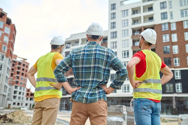 Resultado. Vista posterior de tres hombres con cascos protectores tomados de la mano en la cintura de pie en el sitio de construcción mirando edificios en construcción durante el día