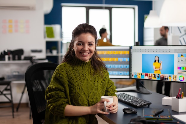 Foto gratuita retocador de mujer mirando a la cámara sonriendo sentado en la agencia de medios creativos retocando fotos de clientes en pc con dos pantallas