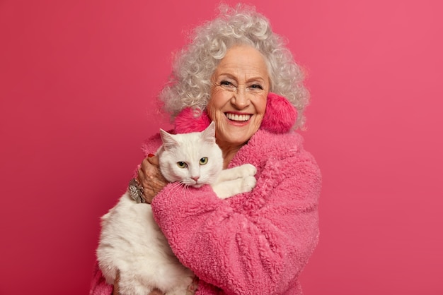 El retrato de la abuela arrugada sostiene el gato blanco en las manos, se queda en casa durante el brote pandémico, usa aretes mullidos, bata suave, va a alimentar a la mascota, aislado en la pared rosa. Mujer, en, pensión