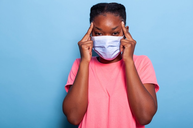 Retrato de adolescente afroamericano pensativo con mascarilla médica protectora contra el coronavirus con los dedos en las sienes. estudiante pensando en la idea de innovación en estudio con fondo azul.