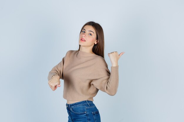 Retrato de una adolescente apuntando hacia la derecha con los pulgares en suéter, jeans y mirando inteligente vista frontal