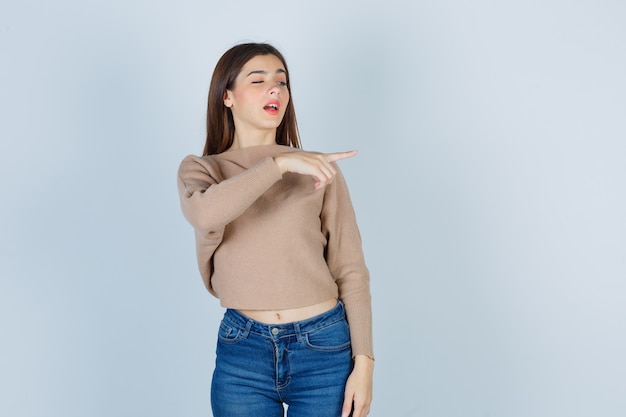 Retrato de una adolescente apuntando a la derecha en suéter, jeans y mirando confiada vista frontal