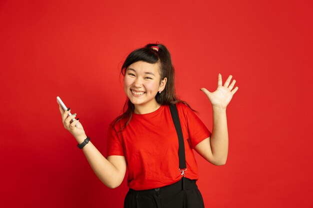 Retrato de adolescente asiático aislado sobre fondo rojo de estudio. Modelo morena mujer hermosa en estilo casual. Concepto de emociones humanas, expresión facial, ventas, publicidad. Feliz, sosteniendo el teléfono inteligente.