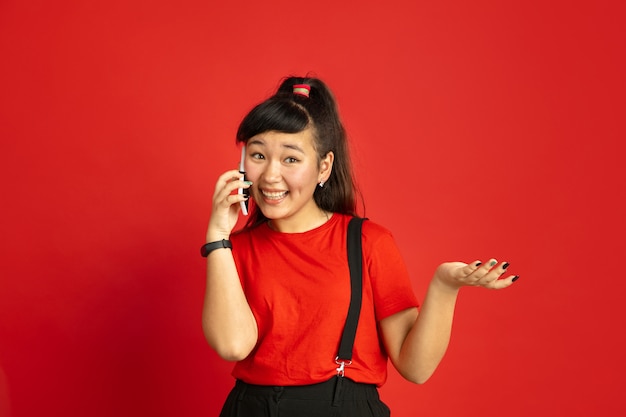 Retrato de adolescente asiático aislado sobre fondo rojo de estudio. Modelo morena mujer hermosa con el pelo largo en estilo casual. Concepto de emociones humanas, expresión facial, ventas, publicidad. Hablando por telefono.