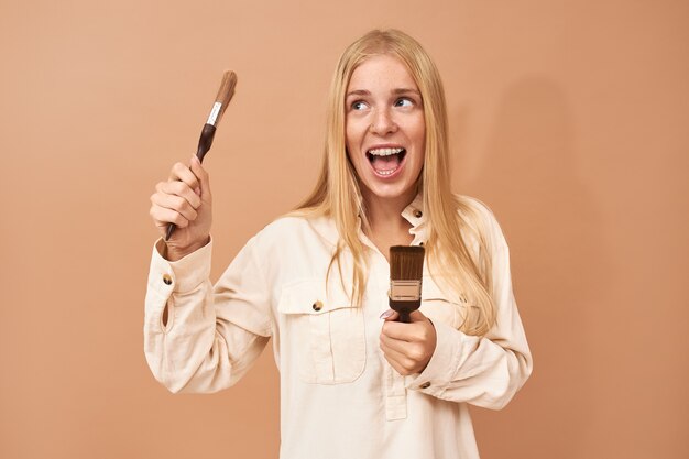 Retrato de una adolescente bonita con tirantes y cabello largo usando herramientas especiales mientras pinta las paredes interiores para protegerlas de daños por agua o corrosión
