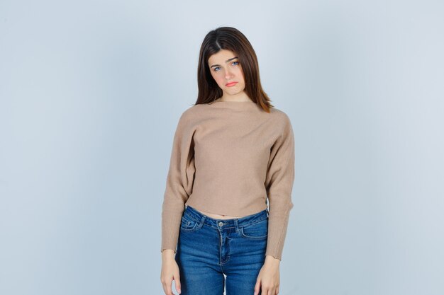 Retrato de una adolescente curvándose el labio inferior en suéter, jeans y mirando decepcionado vista frontal