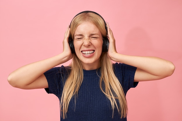 Retrato de una adolescente elegante con un piercing en la nariz manteniendo los ojos cerrados y haciendo muecas escuchando música rock demasiado fuerte en auriculares inalámbricos