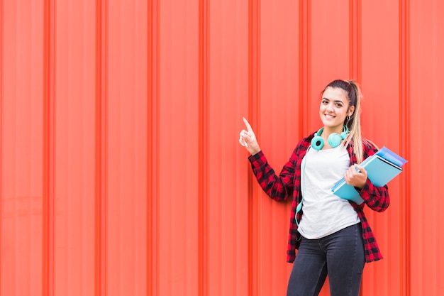 Retrato de una adolescente feliz sosteniendo libros en la mano con auriculares alrededor de su cuello apuntando el dedo contra una pared naranja