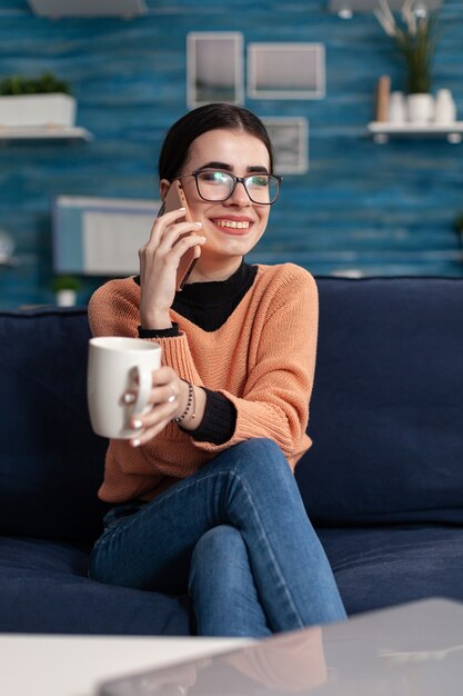 Retrato de adolescente hablando sobre el estilo de vida en el teléfono inteligente moderno mientras se ríe con su amiga sentada en el sofá en la sala de estar. Mujer joven divirtiéndose durante la llamada de conversación de estilo de vida divertido