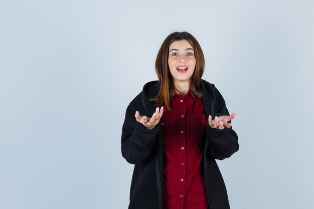 Retrato de una adolescente haciendo gesto de pregunta en camisa, abrigo y mirando perplejo vista frontal