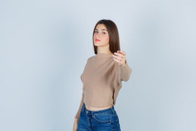Retrato de una adolescente invitando a venir en suéter, jeans y mirando confiada vista frontal