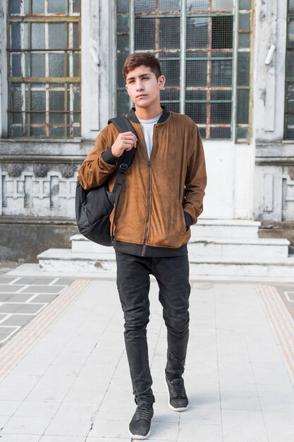 Retrato de un adolescente con la mano en el bolsillo que lleva la bolsa en el hombro caminando frente al edificio