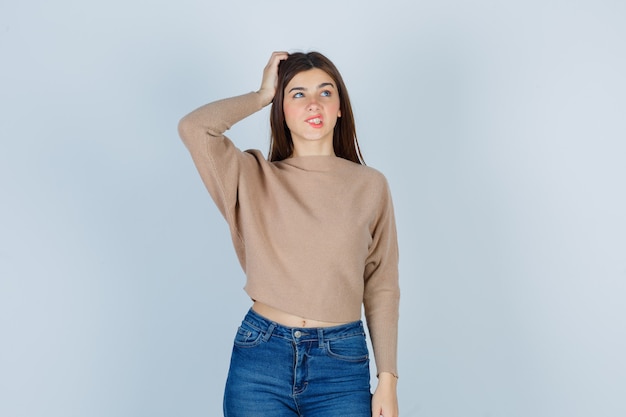 Retrato de una adolescente manteniendo la mano en la cabeza, mordiendo el labio, mirando hacia arriba en suéter, jeans y mirando pensativo vista frontal