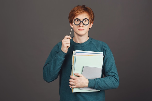 Retrato de adolescente pelirroja guapo divertido en suéter verde de moda con cuadernos en la mano, mirando con los ojos aparecidos a través de vasos de papel en el palo.