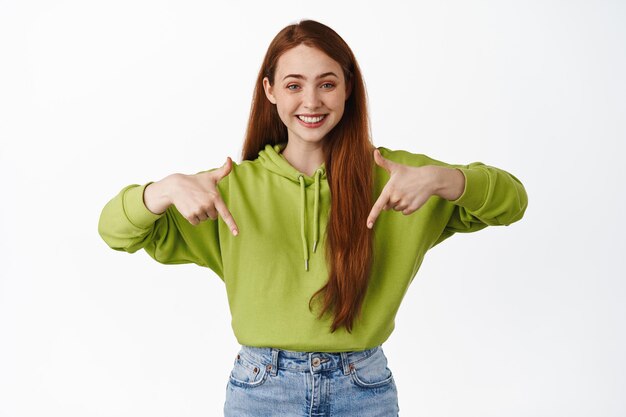 Retrato de una adolescente pelirroja sonriendo con dientes blancos, señalando con el dedo hacia abajo en un descuento promocional, mostrando un anuncio, de pie contra un fondo blanco.