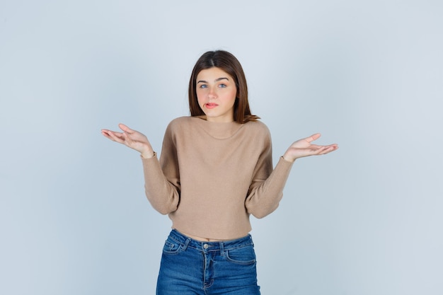 Retrato de una adolescente que muestra un gesto de impotencia en suéter, jeans y mirando desorientado vista frontal