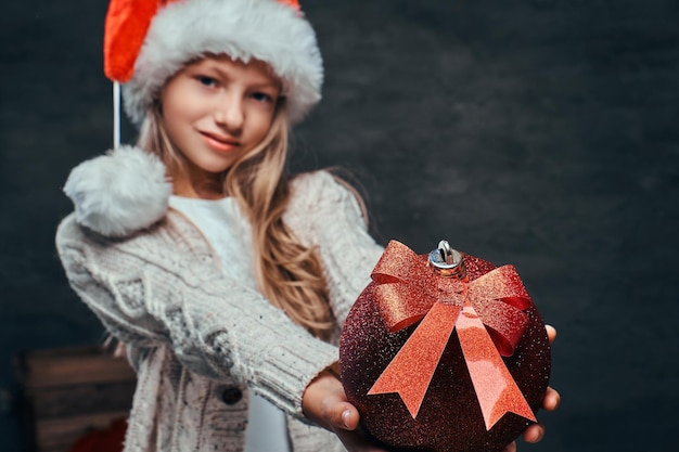 Retrato de un adolescente con sombrero de Papá Noel sosteniendo una gran bola de Navidad sobre un fondo de textura oscura.