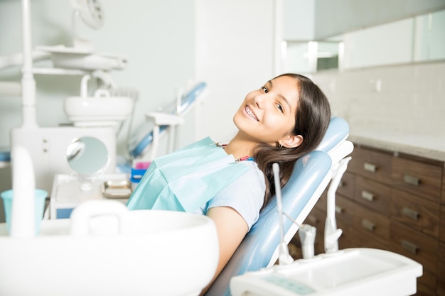 Retrato de una adolescente sonriente con aparatos ortopédicos sentados en una silla en la clínica dental