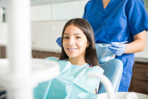 Retrato de una adolescente sonriente con aparatos ortopédicos sentados en una silla mientras el dentista está de pie en la clínica