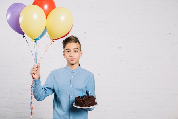 Retrato de adolescente sosteniendo globos y pastel de cumpleaños contra la pared