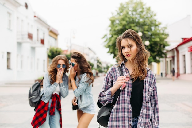 Retrato de una adolescente triste y hermosa con camisa a cuadros con trenzas mirando a la cámara contra dos amigos con gafas de sol y ropa de denim chismorreando sobre ella en la calle. Desenfocado.