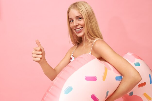 Retrato de adorable alegre joven mujer europea con cabello lacio y rubio que lleva un anillo de natación rosa debajo del brazo, va a nadar en la piscina o el mar, sonriendo