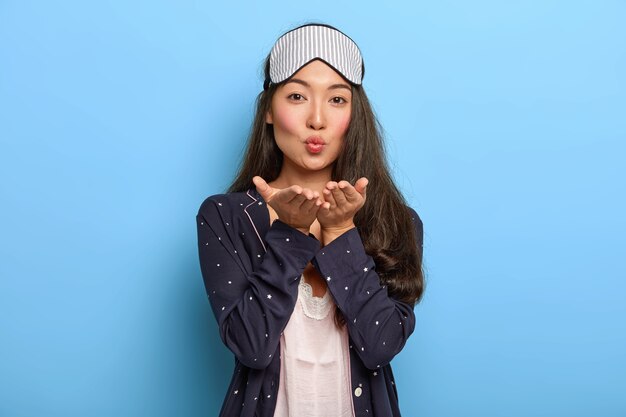 Retrato de adorable mujer morena Asain envía beso de aire a su esposo, se despierta por la mañana, usa máscara para dormir y traje de noche, descansa en casa