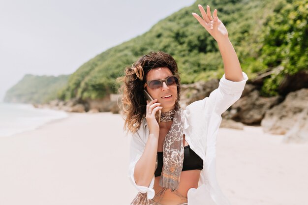 Retrato de adorable mujer romántica vestida con top negro, camisa blanca habla en el teléfono inteligente