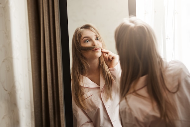 Retrato de adorable mujer rubia juguetona haciendo bigote de mechón de pelo, mirándose en el espejo y haciendo cara graciosa