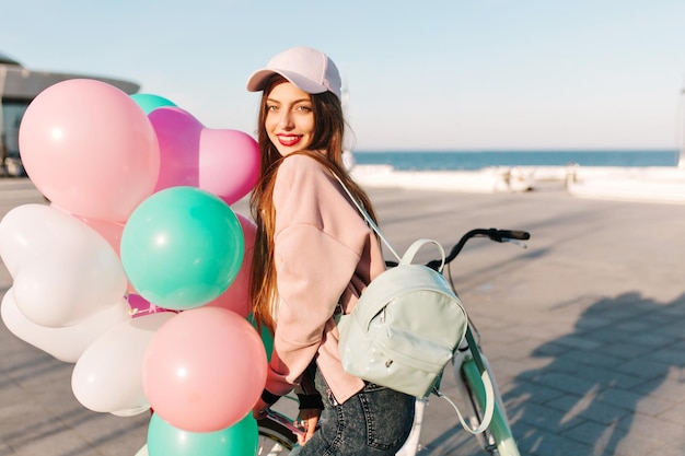 Retrato de adorable niña morena sonriente en gorra rosa de moda con mochila caminando por el muelle del océano con globos de colores. Linda mujer joven con cabello largo posando en el muelle del mar después de la fiesta de cumpleaños