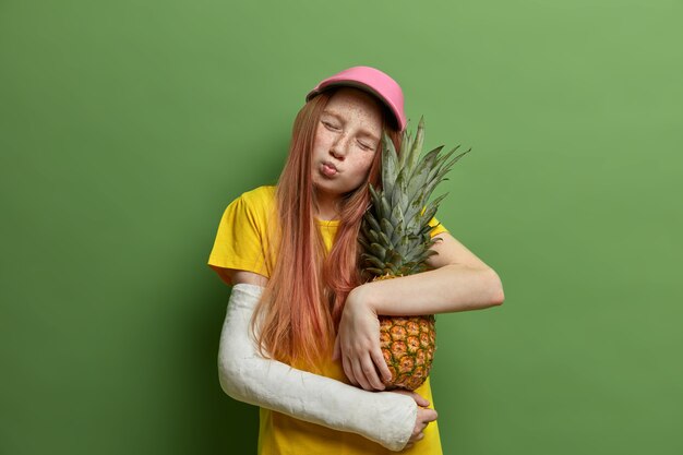 Retrato de adorable niña pecosa inclina la cabeza, tiene los ojos cerrados y los labios redondeados, abraza una deliciosa piña con amor, se ha roto el brazo después de caer de altura, aislado en una pared verde.