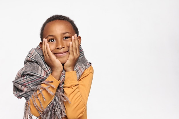 Foto gratuita retrato de adorable niño de piel oscura en abrigo cálido tomados de la mano en las mejillas y mirando hacia arriba con una sonrisa alegre