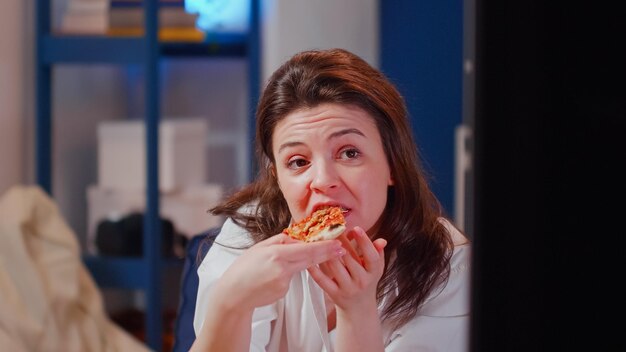 Retrato de un adulto caucásico mordiendo una rebanada de pizza en casa mientras sostiene una botella de cerveza. Mujer joven mirando una película en la televisión y comiendo comida para llevar con comida rápida y bebida alcohólica