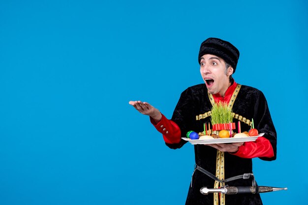 Retrato de un adulto joven sorprendido con vestido tradicional sosteniendo una bandeja llena de dulces nacionales apuntando hacia adelante sobre el fondo de la ola azul