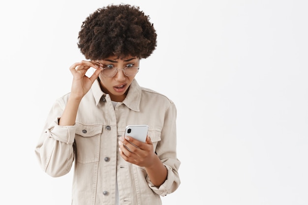 Retrato de afroamericana intensa confundida y cuestionada no puede creer en tonterías que leyó a través de smarpthone, quitándose las gafas, haciendo muecas, mirando con estupor a la pantalla