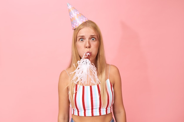 Foto gratuita retrato aislado de una adolescente divertida emocional con cabello rubio recto y sombrero cónico saliendo los ojos después de haber sorprendido la expresión facial debido a la inesperada fiesta de cumpleaños