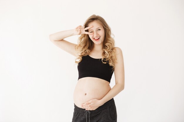 Retrato aislado de alegre joven embarazada europea con cabello ondulado rubio en ropa cómoda mostrando lengua