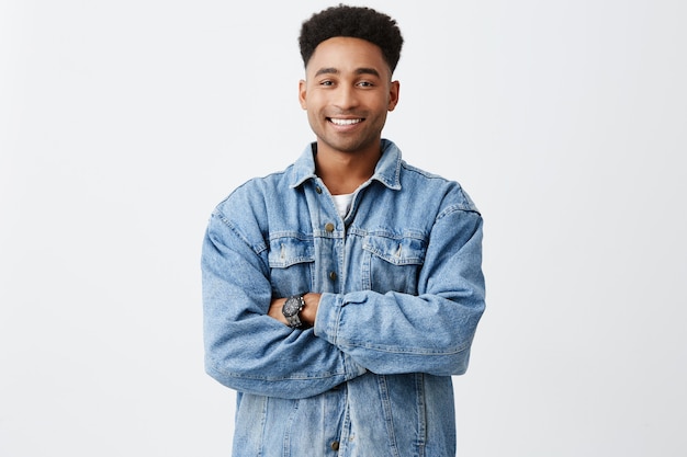 Retrato aislado de joven gracioso hombre de piel oscura con los brazos cruzados con peinado afro en camisa blanca casual debajo de la chaqueta de mezclilla con expresión de la cara emocionada