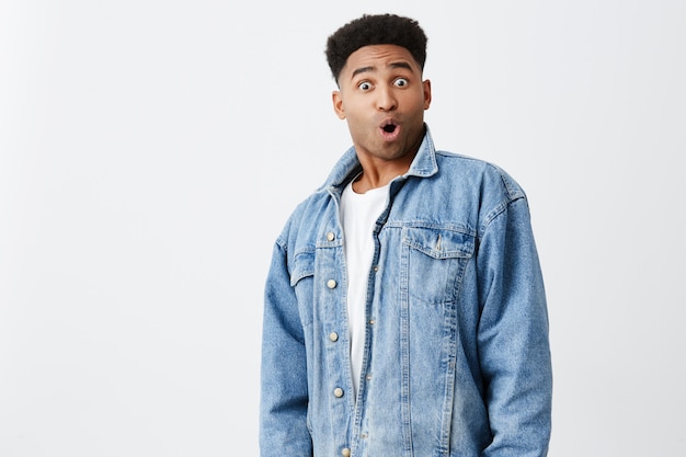 Retrato aislado del joven hombre de piel oscura divertido sorprendido con peinado afro en camisa blanca casual debajo de la chaqueta de mezclilla con expresión de la cara emocionada