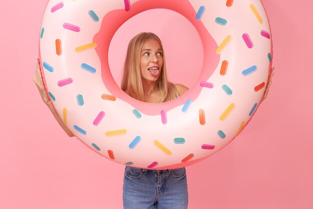 Retrato aislado de moda joven rubia con jeans rotos divirtiéndose durante las vacaciones de verano, sosteniendo un anillo de natación rosa