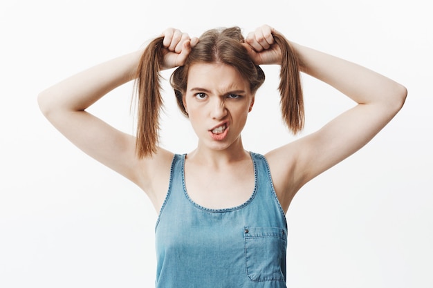 Retrato aislado de la mujer caucásica joven divertida con el pelo oscuro en camisa azul brillante que sostiene el pelo con las manos, con expresión tonta, mostrando los dientes.