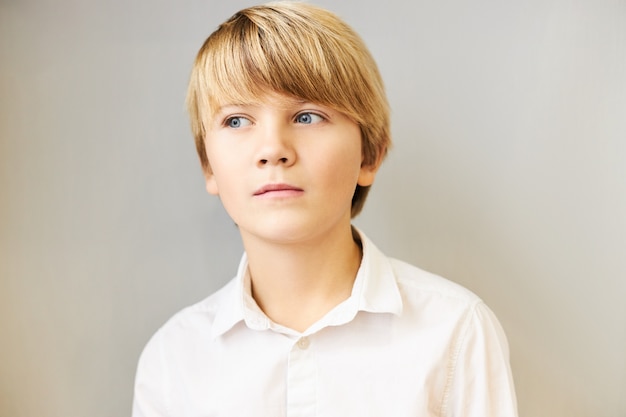 Retrato aislado de un niño caucásico asombrado con flecos y ojos azules mirando a otro lado con misteriosa expresión pensativa, sumido en pensamientos, reflexionando, teniendo una idea o haciendo un plan, posando en la pared en blanco