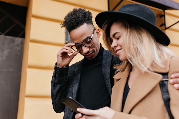 Retrato al aire libre de alegre niña caucásica mostrando nuevo teléfono para amigo africano. Elegante joven negro con gafas divirtiéndose con una mujer rubia en las calles de la ciudad.