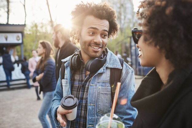 Retrato al aire libre de amigos afroamericanos caminando en el parque mientras habla y toma café, vistiendo ropa de moda.