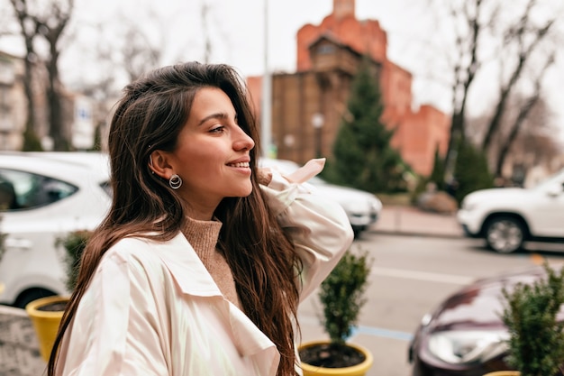 Retrato al aire libre de una atractiva mujer europea con estilo con el pelo oscuro con bata blanca caminando por la ciudad en primavera de cerca