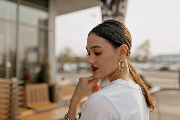 El retrato al aire libre de una atractiva mujer morena con labios rojos lleva ganancias doradas y una camiseta blanca