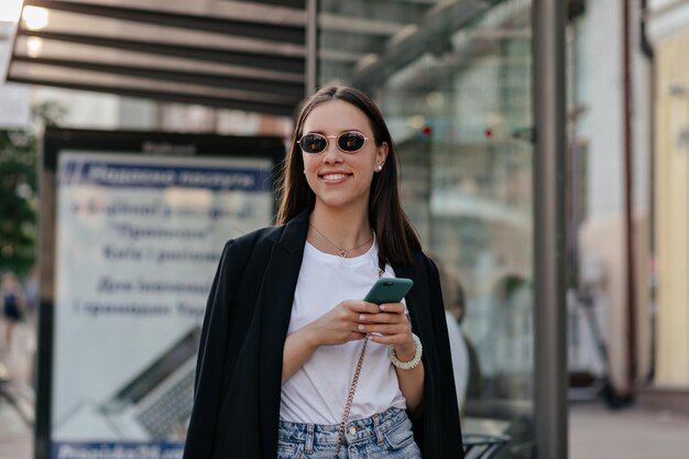 Retrato al aire libre de una chica bastante elegante con chaqueta oscura y gafas negras está desplazando el teléfono inteligente y sonriendo mientras camina por la ciudad en un día soleado y cálido